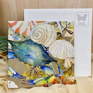 May include: A watercolor painting of a blue crab with seashells and seaweed on a brown background. The painting is on a card with a white envelope.