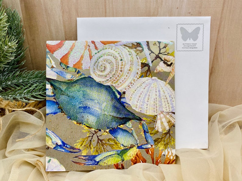 May include: A watercolor painting of a blue crab with seashells and seaweed on a brown background. The painting is on a card with a white envelope.