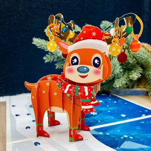 May include: A 3D pop-up card featuring a reindeer wearing a red and white Santa hat and a red and green scarf. The reindeer has a brown nose and large, brown eyes. The card is decorated with snowflakes and a blue background.