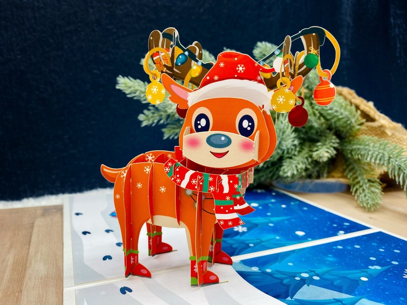 May include: A 3D pop-up card featuring a reindeer wearing a red and white Santa hat and a red and green scarf. The reindeer has a brown nose and large, brown eyes. The card is decorated with snowflakes and a blue background.