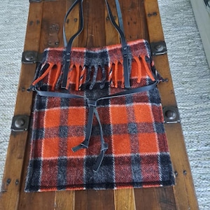 May include: A plaid tote bag with black leather straps and a matching belt. The bag features a fringed top and a pattern of orange, black, and gray squares. The bag is sitting on a wooden surface.