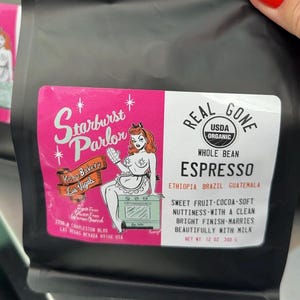 May include: Black bag of Real Gone Espresso whole bean coffee. The pink label features a retro illustration of a woman and the text "Starburst Parlor Keto Bakery Las Vegas". The bag contains 12 oz (340 g) of coffee.