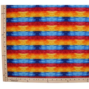 May include: A fabric featuring horizontal stripes in repeating patterns of blue, red, orange, and yellow. A wooden ruler is positioned along the left and bottom edges, showing measurements in inches up to 24 inches along the side and 24 inches along the bottom.