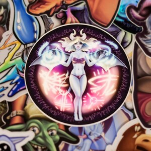 May include: A round sticker with a black background and a white female figure with wings. The figure is surrounded by a glowing pink and blue aura. The sticker has a black border with a repeating pattern.