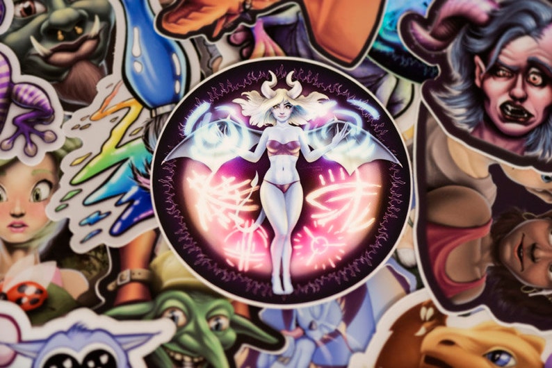 May include: A round sticker with a black background and a white female figure with wings. The figure is surrounded by a glowing pink and blue aura. The sticker has a black border with a repeating pattern.