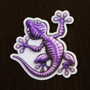 May include: A purple and white gecko sticker with a striped pattern. The gecko is in a climbing pose with its tail curled up.