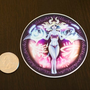 May include: A round sticker with a purple, pink, and blue design. A female figure with horns and wings is in the center of the sticker. The figure is wearing a pink and white outfit.