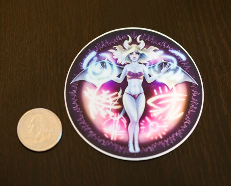 May include: A round sticker with a purple, pink, and blue design. A female figure with horns and wings is in the center of the sticker. The figure is wearing a pink and white outfit.