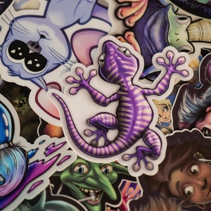 K&ouml;nnte beinhalten: Eine Sammlung bunter Aufkleber mit verschiedenen Designs, darunter ein lila Gecko, ein blauer Pinsel, eine Katze und ein Kobold. Die Aufkleber sind auf wei&szlig;em Papier gedruckt und haben eine gl&auml;nzende Oberfl&auml;che.