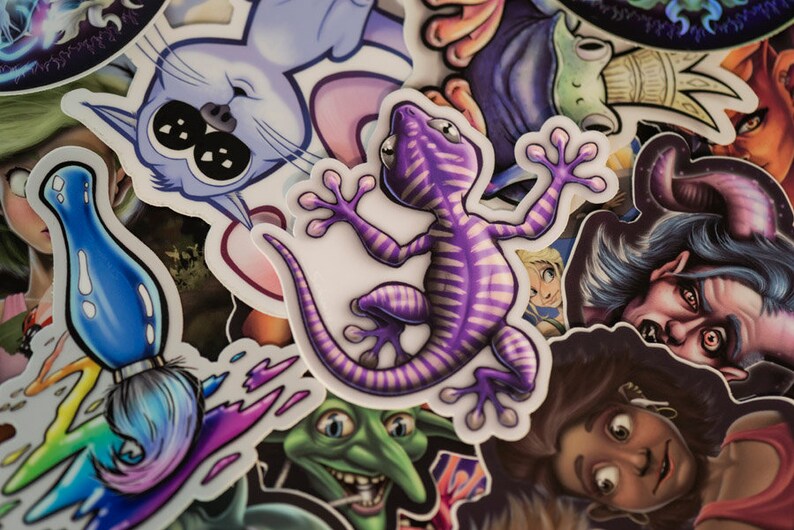 K&ouml;nnte beinhalten: Eine Sammlung bunter Aufkleber mit verschiedenen Designs, darunter ein lila Gecko, ein blauer Pinsel, eine Katze und ein Kobold. Die Aufkleber sind auf wei&szlig;em Papier gedruckt und haben eine gl&auml;nzende Oberfl&auml;che.