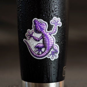 K&ouml;nnte beinhalten: Ein schwarzer Edelstahlbecher mit einem violetten Gecko-Aufkleber. Der Becher ist mit Kondensationstropfen bedeckt.