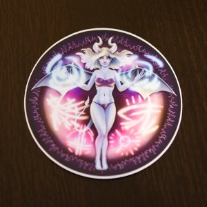 May include: A round sticker featuring a digital illustration of a woman with horns and wings. She is wearing a red bikini top and a purple skirt. The background is a dark purple with glowing pink and white accents.