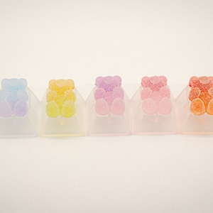 Könnte beinhalten: Sieben transparente Kunststoff-Tastenkappen mit Gummibärchen-Designs in einem Regenbogen-Farbverlauf. Die Tastenkappen sind in einer Reihe von links nach rechts angeordnet, beginnend mit Hellblau und endend mit Orange.