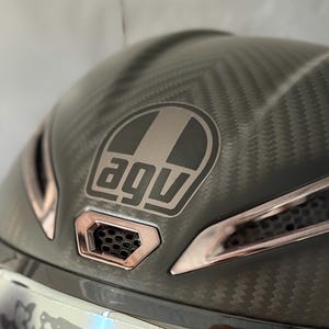 May include: A close-up of a motorcycle helmet with a carbon fibre pattern. The helmet features the "AGV" logo in a rectangular design, with bronze-coloured accents. The visor is silver with a speckled design.