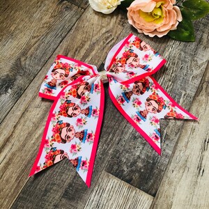 May include: A large white and pink bow with a pink trim. The bow is decorated with a repeating pattern of a woman's face with flowers. The bow has a rhinestone in the center.