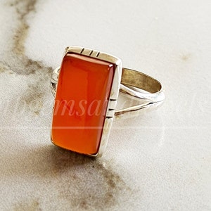 Könnte beinhalten: Ein silberner Ring mit einem orangefarbenen Karneol-Stein. Der Stein ist rechteckig und hat eine glatte, polierte Oberfläche.