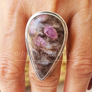 Può includere: Un anello d'argento con una pietra preziosa rosa e marrone a forma di lacrima. L'anello è su un dito.