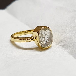 May include: A gold ring featuring a large, clear, square-cut gemstone. The ring's band has a textured design, and the setting is a simple bezel. The ring is displayed against a white backdrop, accentuating the gold and the gemstone's clarity.