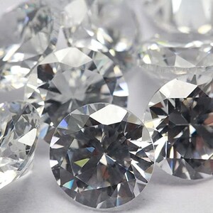 May include: A group of clear, round, faceted gemstones, likely diamonds or cubic zirconia, on a white surface.