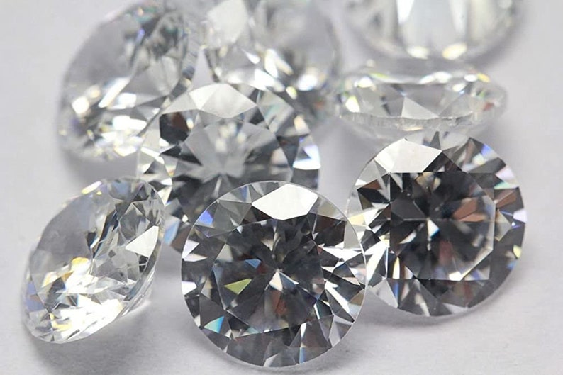 May include: A group of clear, round, faceted gemstones, likely diamonds or cubic zirconia, on a white surface.