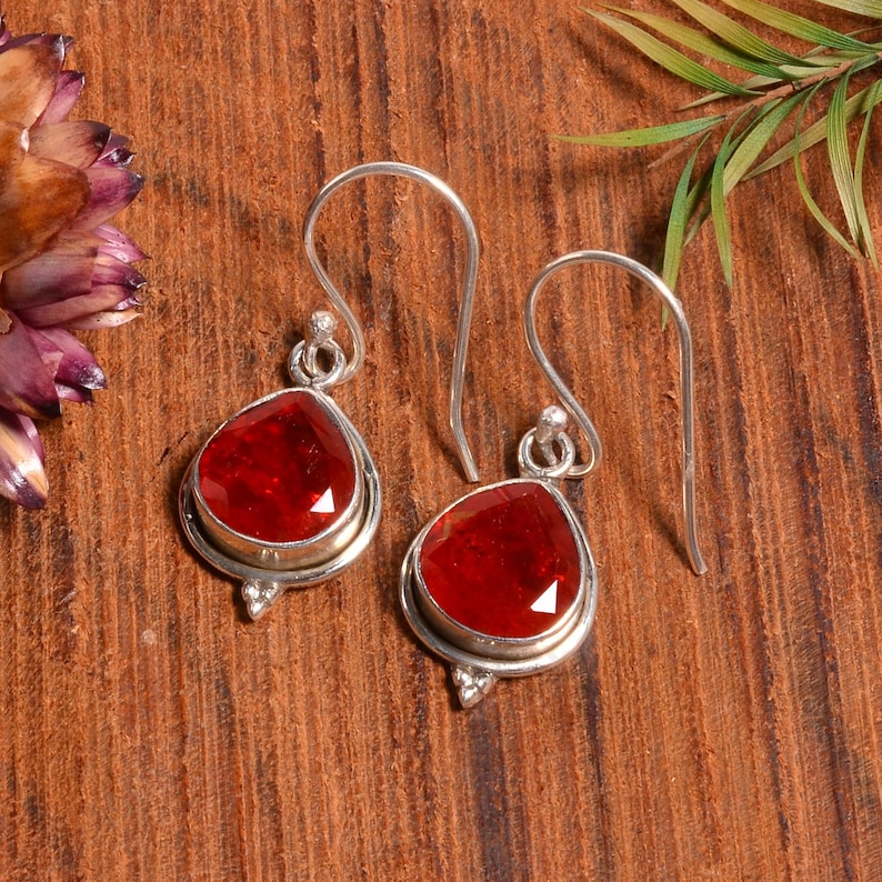 Peut inclure: Une paire de boucles d'oreilles pendantes en argent avec des gouttes de pierres pr&eacute;cieuses rouges. Les boucles d'oreilles ont un design simple avec une petite boucle en argent en haut et une plus grande boucle en argent en bas. Les pierres pr&eacute;cieuses rouges sont facett&eacute;es et ont une forme de larme.