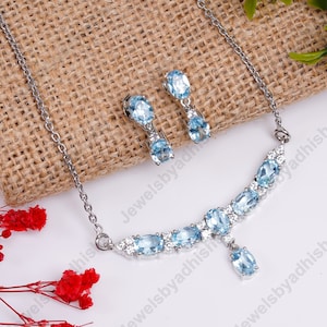 May include: A silver necklace and earrings set with a light blue gemstone pendant and matching earrings. The necklace features a curved design with a row of smaller gemstones.