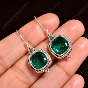 Può includere: Un paio di orecchini pendenti in argento con pietre preziose verde smeraldo quadrate. Le pietre sono incastonate in un bordo decorativo a forma di corda. Design a gancio semplice. Sfondo scuro.