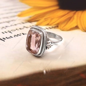 May include: A silver ring with a pink gemstone set in a square bezel. The ring has a detailed band with a rope-like design and a small floral accent.