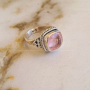 May include: A silver ring with a pink gemstone set in a square shape. The ring has a rope-like design around the band and a small cluster of beads on one side.
