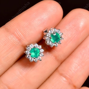 May include: A pair of stud earrings featuring a central emerald-green gemstone surrounded by a halo of smaller, clear gemstones. The earrings are set in a silver-colored metal, creating a floral-like design. The earrings are displayed on a light skin tone.