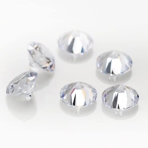 May include: Six clear round faceted gemstones, likely diamonds, scattered on a white surface.