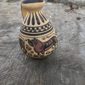 May include: A small, antique-style ceramic jug with a handle. The jug features black and red painted designs on a beige background. The design includes a stylized animal figure and geometric patterns. The top and handle are dark brown.
