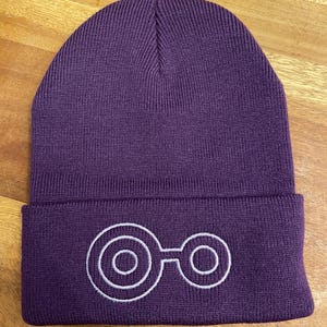 Könnte beinhalten: Lilafarbener Strickmütze mit einem weißen gestickten Symbol aus zwei ineinandergreifenden Kreisen.