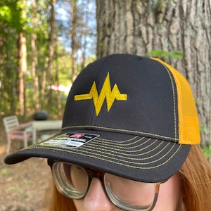 May include: A black and gold baseball cap with a yellow embroidered heartbeat logo. The cap has a mesh back and a black adjustable strap.