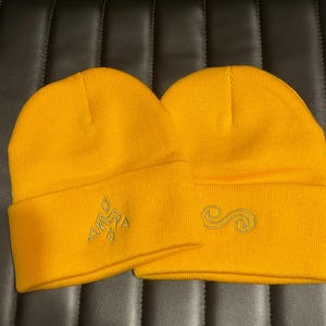 May include: Two yellow knit beanies with different embroidered designs. One beanie has a geometric design and the other has a swirl design.