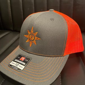 Puede incluir: Gorra de camionero gris y naranja con un diseño de sol naranja bordado. La gorra tiene una visera curva y una correa ajustable. La etiqueta de la marca Richardson es visible en la parte delantera.