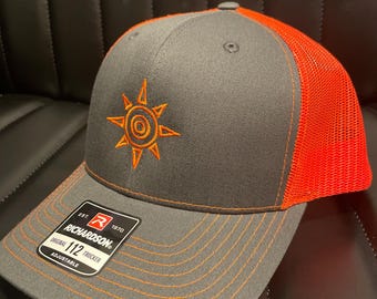 Gorra de béisbol bordada con el Escudo del Valor de Digimon Adventure, estilo camionero, con malla. Color carbón y naranja con hilo naranja.