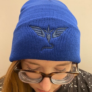 Può includere: Un berretto blu reale con un logo ricamato blu navy di un uccello con le ali spiegate.