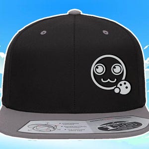May include: A black and gray baseball cap with a white cartoon face and a white circle with a black dot inside. The cap has a gray brim and a white label with the text "FLEXFIT ONE SIZE" on the front.