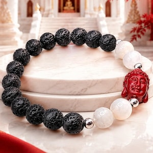 Può includere: Un braccialetto di perline composto da una serie di perle di pietra lavica nera porosa, perle bianche lisce e traslucide e una singola perla rossa intagliata a forma di testa di Buddha. Piccoli distanziatori metallici argentati separano le perle.
