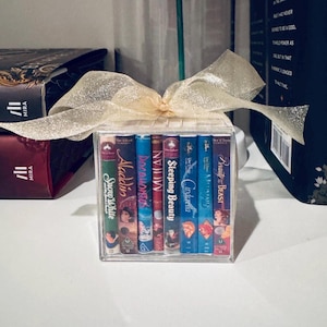 May include: A clear acrylic box tied with a gold ribbon, containing a collection of Disney VHS tapes. Titles include "Snow White," "Aladdin," "Mulan," "Sleeping Beauty," "Cinderella," and "Beauty and the Beast."