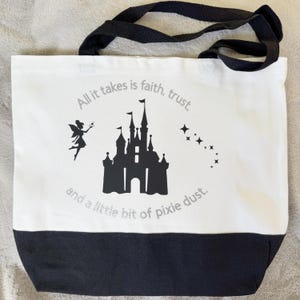 May include: White canvas tote bag with black accents and handles. Features a black silhouette of a castle, a fairy, and text that reads "All it takes is faith, trust, and a little bit of pixie dust."