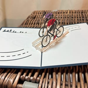 May include: A pop-up card featuring two cyclists racing on a road. The cyclists are wearing purple and red jerseys. The card is white with black road markings and a blue border. The card is open and displayed on a wicker basket.