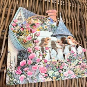 May include: A decorative papercraft scene featuring a quaint house with a blue roof and a chimney. The house is surrounded by a white picket fence and vibrant pink and white flowers. Three small birds perch on the fence.