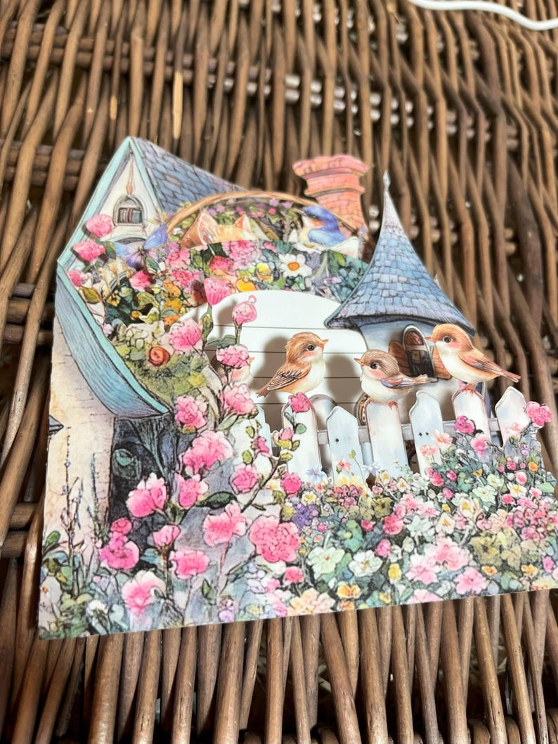 May include: A decorative papercraft scene featuring a quaint house with a blue roof and a chimney. The house is surrounded by a white picket fence and vibrant pink and white flowers. Three small birds perch on the fence.