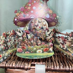 May include: A charming pop-up card depicting a fairy garden. The card includes a mushroom-shaped structure, a hot air balloon, and various woodland creatures such as fairies, a rabbit, and a bee, set amongst colourful flowers and butterflies.