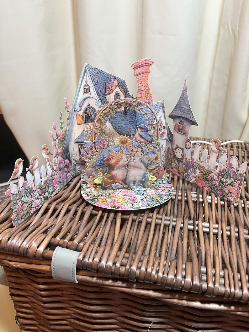 May include: A whimsical 3D paper art scene featuring a cottage, a tower, and a floral archway. Two kittens are nestled in a garden, surrounded by flowers and birds. The scene is set on a woven basket, creating a charming, storybook aesthetic.