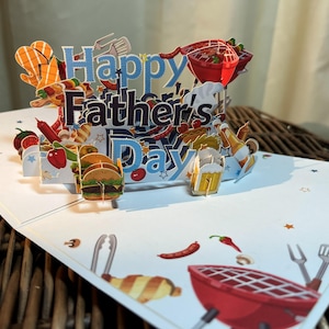 May include: A Father's Day pop-up card with a barbecue theme. The card displays the words "Happy Father's Day" in blue and black. The card includes images of a grill, burgers, beer, and other barbecue-related items.