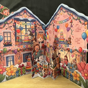 May include: A pop-up birthday greeting card featuring a pink house illustration with colorful balloons, gift boxes, and a rainbow cake. The text "HAPPY BIRTHDAY" is displayed on banners. An elf sits on the roof, and various characters celebrate.