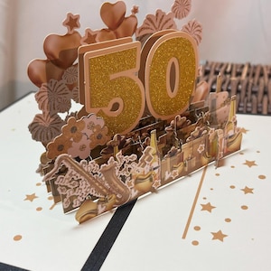 Puede incluir: Una tarjeta de cumpleaños emergente con un "50" dorado con purpurina en el centro. La tarjeta presenta elementos decorativos como corazones, estrellas e instrumentos musicales. La tarjeta está sobre una superficie blanca con detalles de estrellas doradas.
